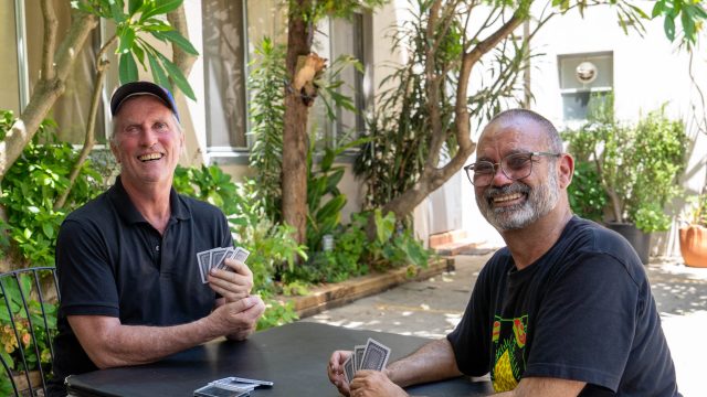 Two men sittong in a courtyard playing cards. they are looking at the camera and smiling.