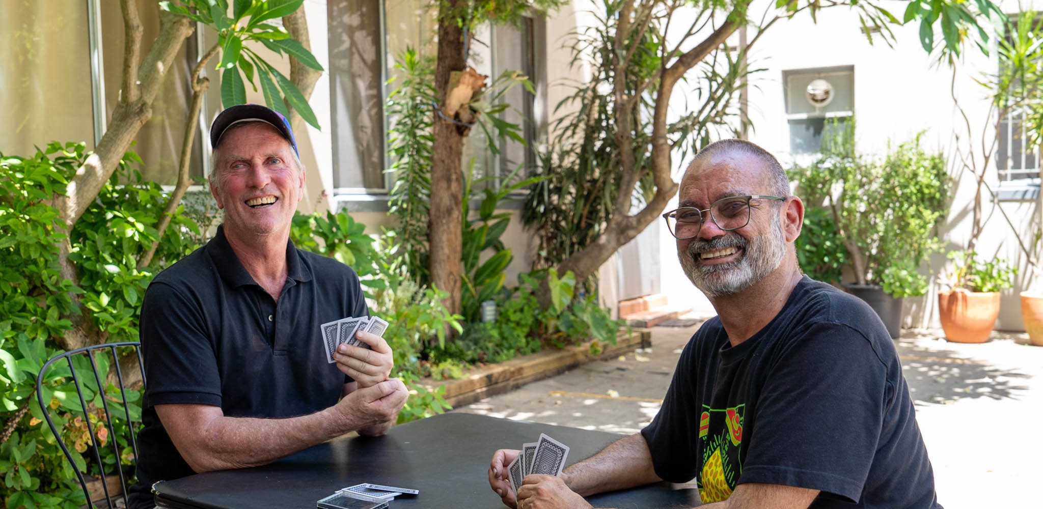 Two men sittong in a courtyard playing cards. they are looking at the camera and smiling.